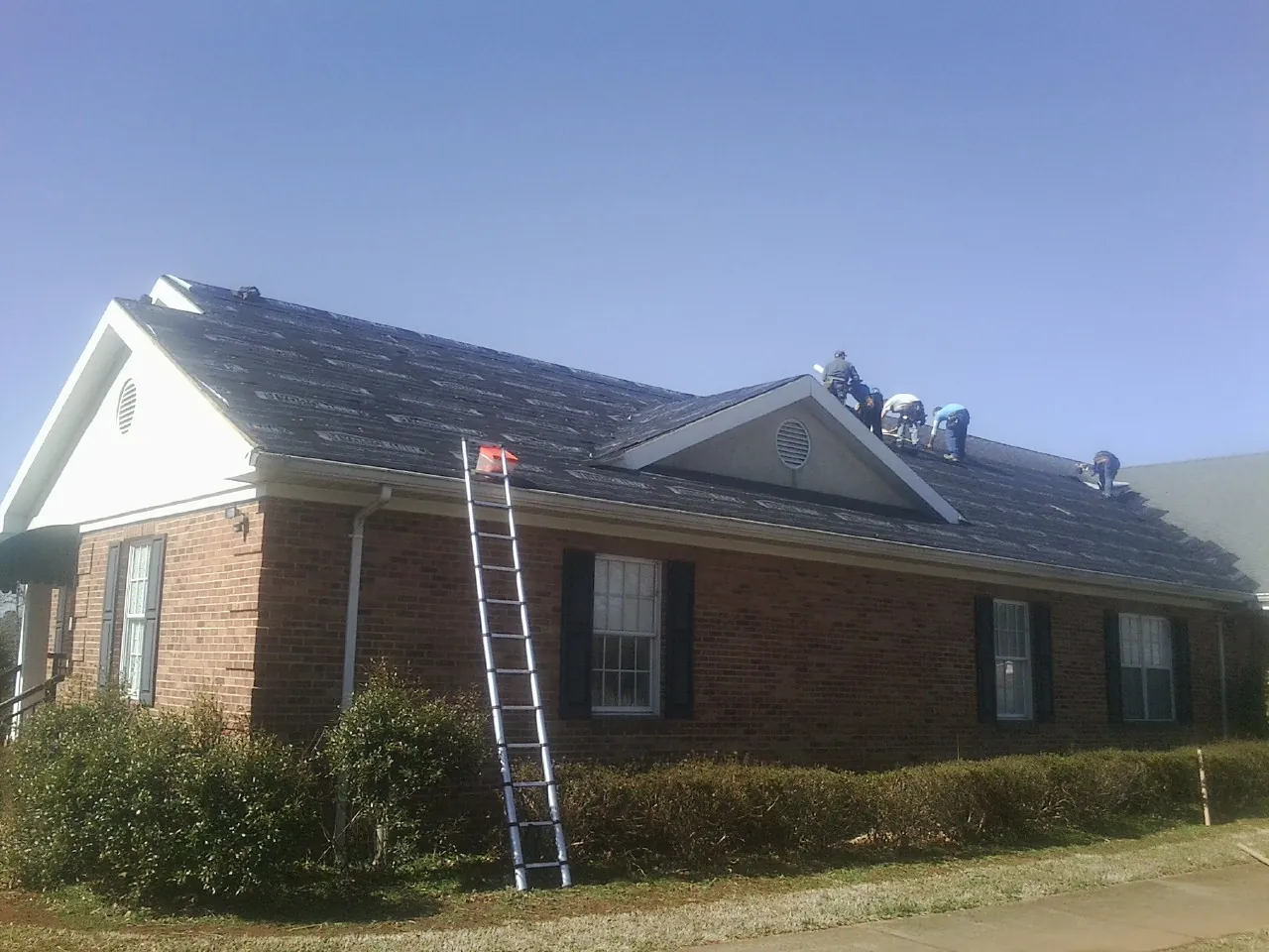 Crew tearing shingles off a brick ranch roof with a ladder set — Emergency Roof Repair work by our crew in Vernon